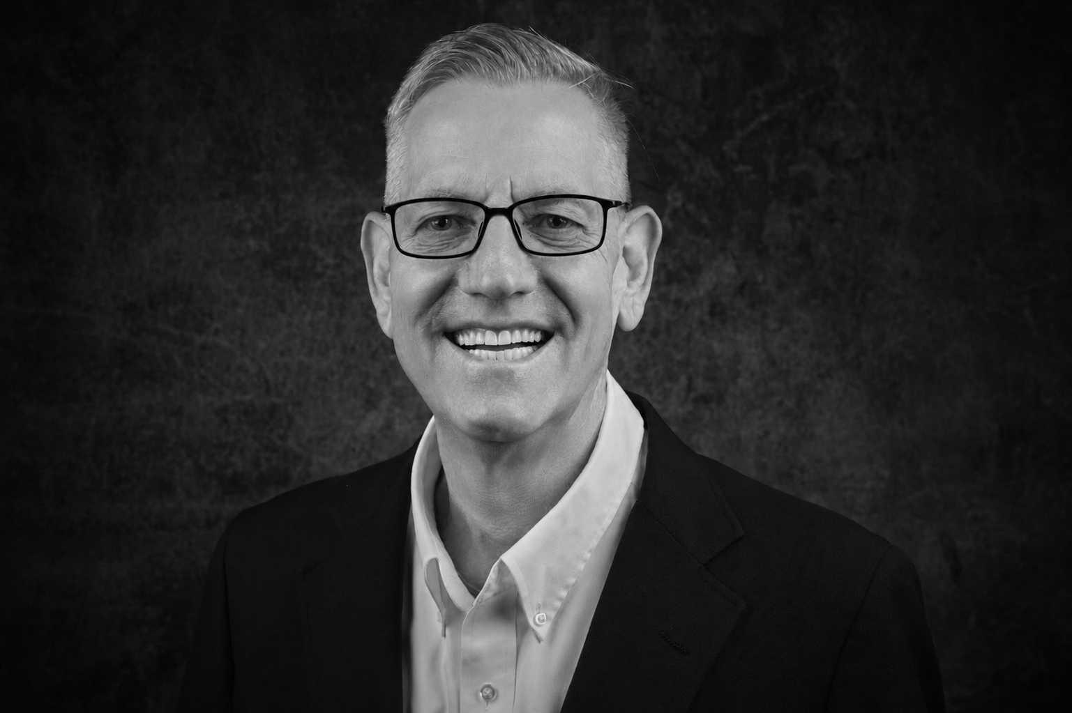 Smiling man with short gray hair and glasses in a dark blazer, portrait-style headshot (black and white).